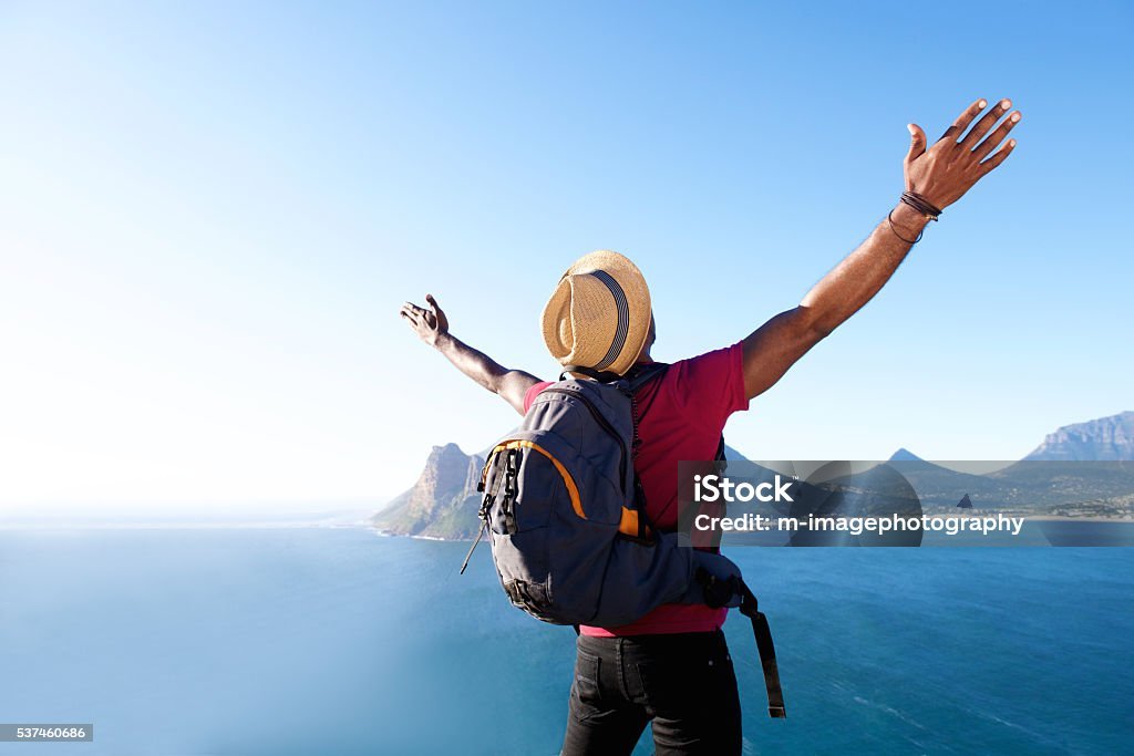 Rear view portrait of young african guy with hat and backpack standing outdoors with arms spread open
